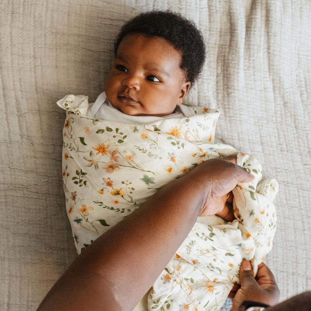 Baby wrapped in a floral swaddle blanket with an adult's hand adjusting it, on a textured fabric background.