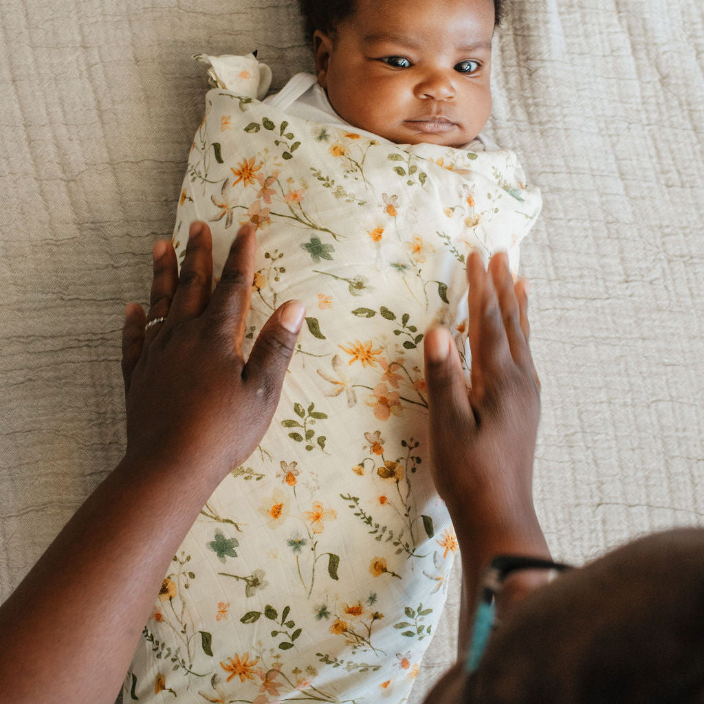 Baby swaddled in a floral blanket with adult hands on either side, lying on a textured surface.