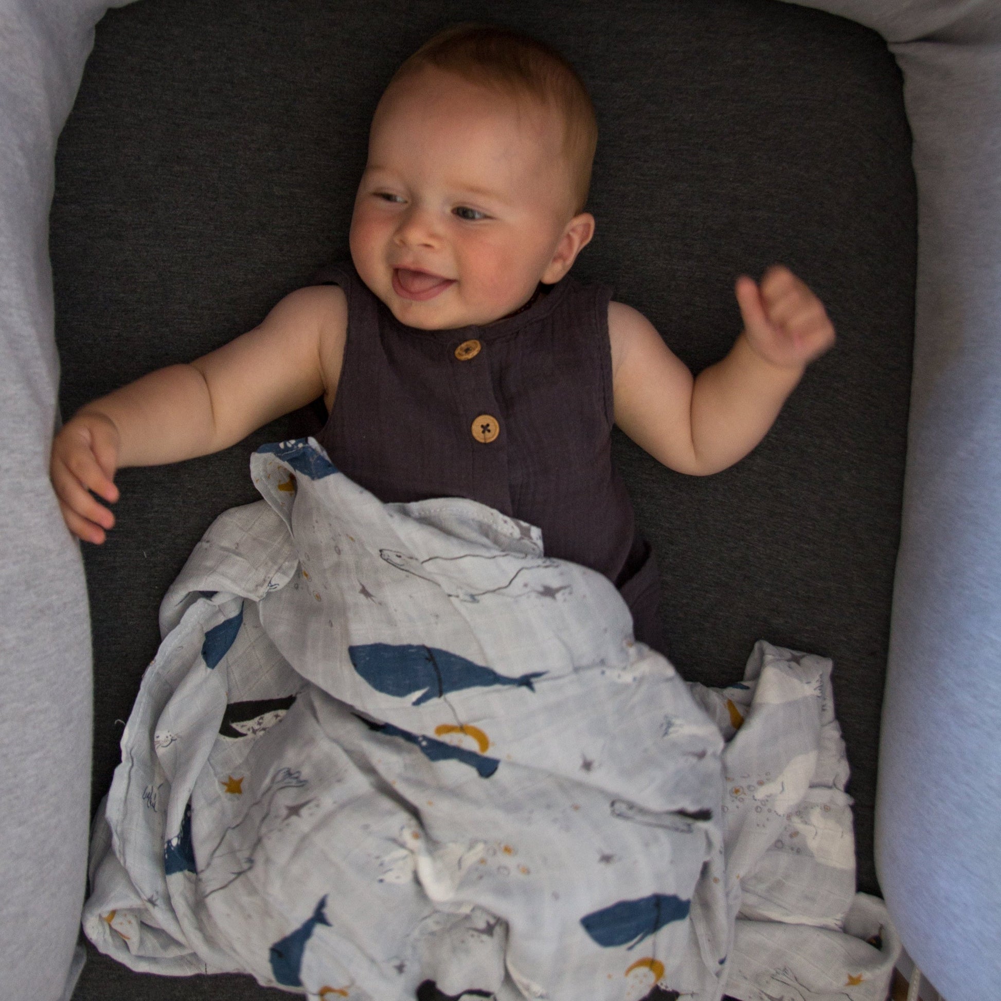 Baby lying in a crib with a blanket, smiling.