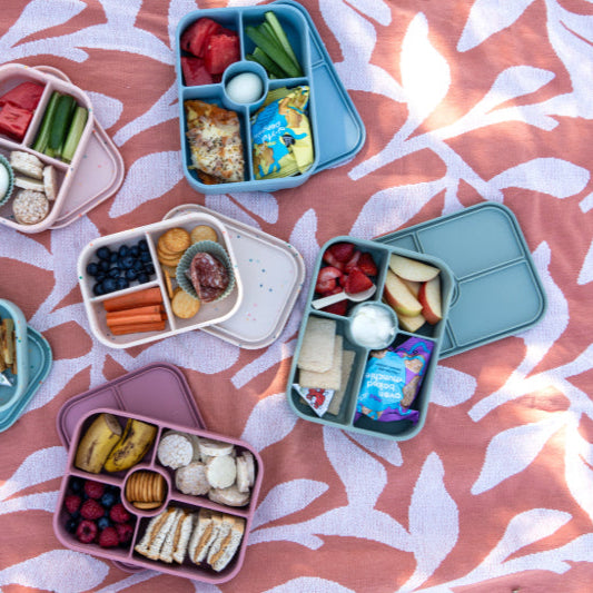 Collection of colorful bento boxes with snacks on a pink leaf-patterned surface