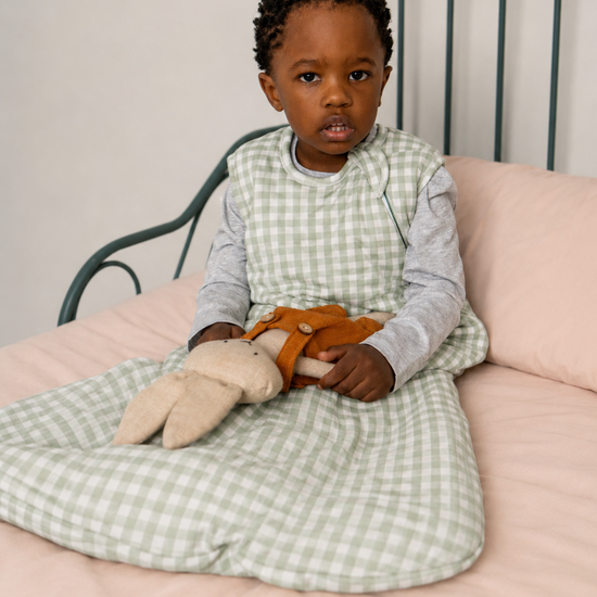 Child sitting on a bed holding teddy bears, wearing a green checkered sleep sack