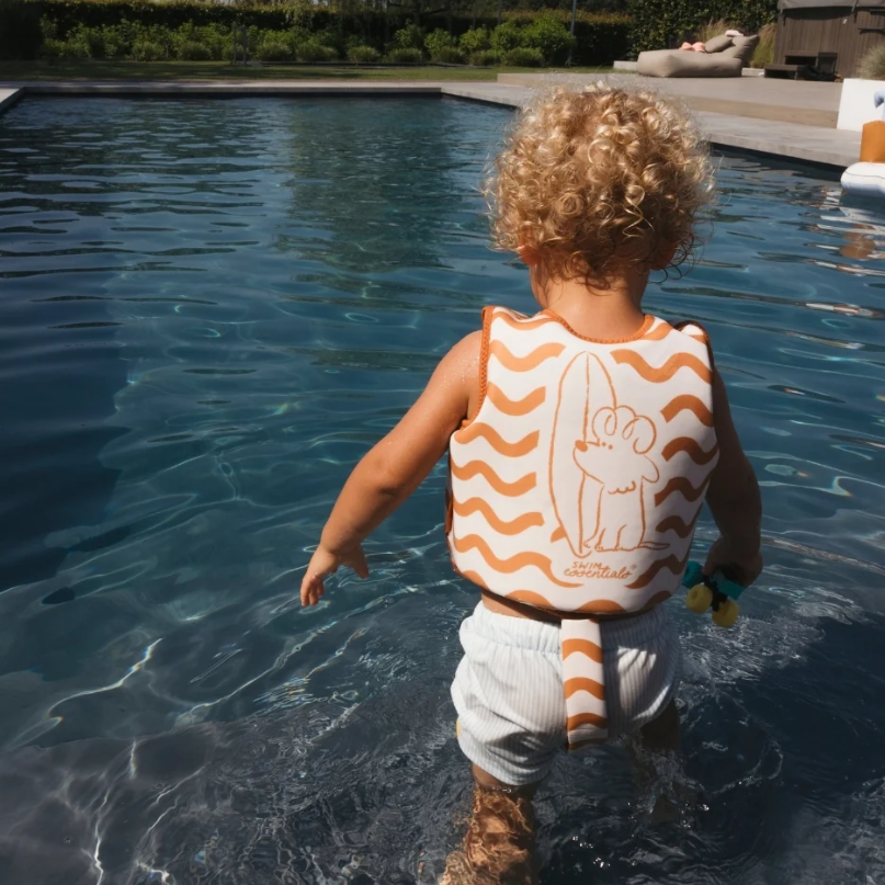 A toddler with curly blonde hair stands in a swimming pool wearing an orange and white wavy-patterned life vest with a cartoon bear design on the back, paired with white swim trunks.