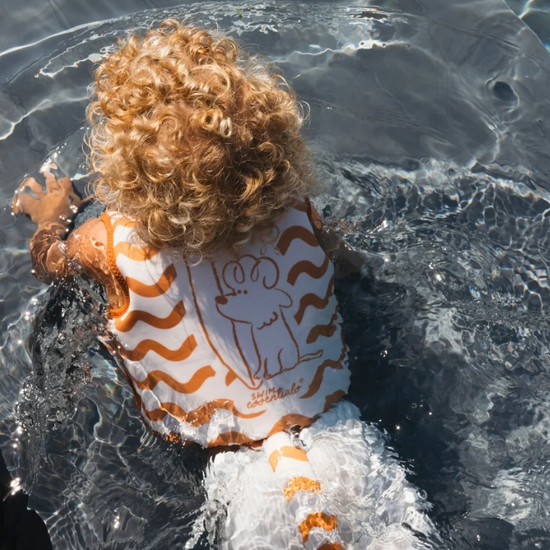 A child with curly blonde hair wearing a white and orange striped swim vest with a dog outline, swimming in clear water.