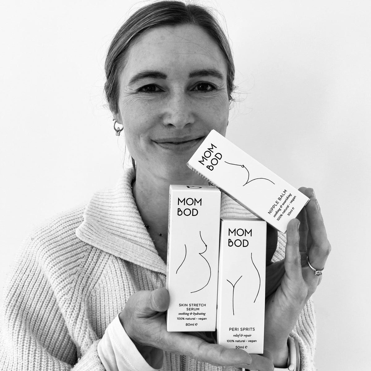A woman in a light-colored knit sweater holds three white boxes of MOM BOD stretch products against a plain white background.