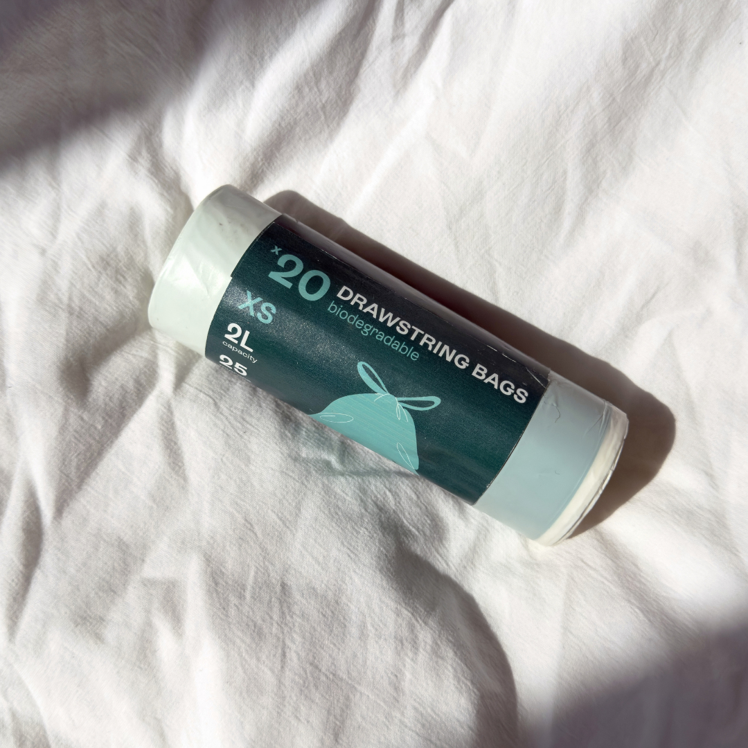 A roll of 20 drawstring bags, 2L each, in a dark teal and white container, placed on a wrinkled white fabric surface with strong natural shadows.
